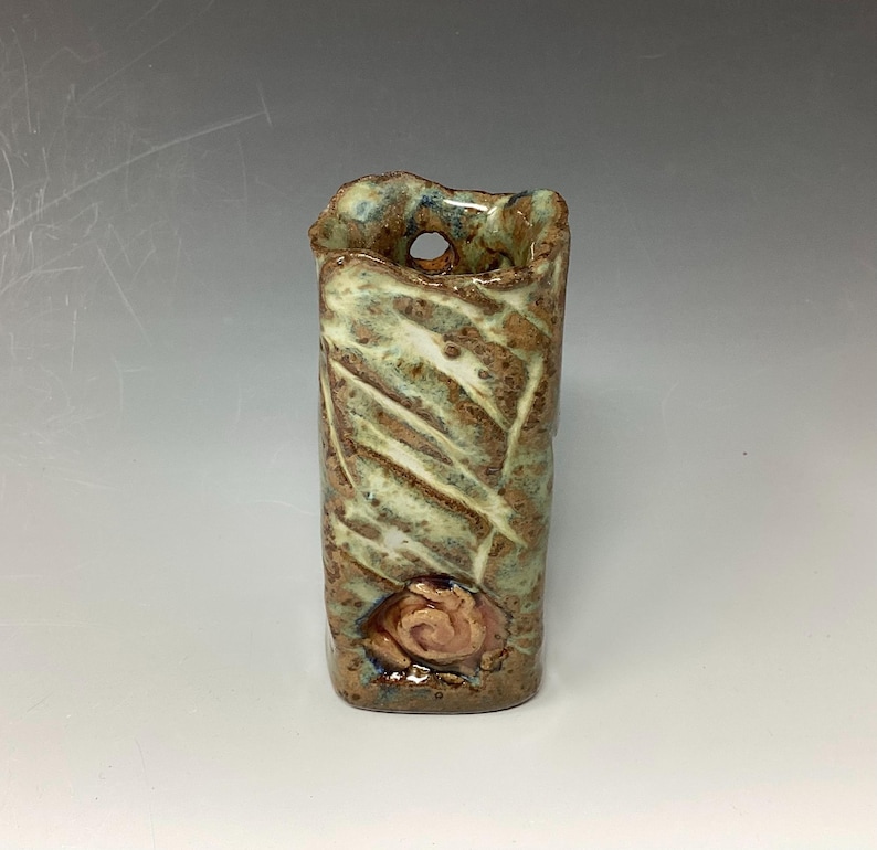 May include: A small, green and brown ceramic vase with a textured surface and a small hole near the top. The vase has a decorative design on the front.