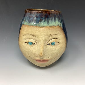 Puede incluir: Un jarrón de cerámica con forma de cara con ojos azules y un esmalte marrón claro.