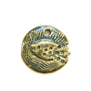 May include: A green and gold ceramic pendant with a raised fish design. The fish has a textured body and a hole for hanging.