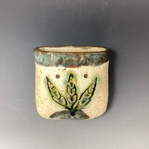 以下が含まれることがあります： 緑と茶色の葉のデザインが施された、薄い白の小さな陶器の花瓶。花瓶は薄い青色の釉薬がかけられた縁をしています。