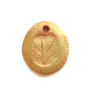 May include: A small, oval-shaped ceramic pendant with a light brown glaze and a raised leaf design. The pendant has a small hole at the top for hanging.