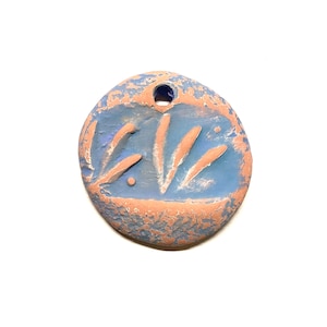 May include: A round, blue and pink clay pendant with a hole at the top for hanging. The pendant has a design of lines and dots carved into the surface.