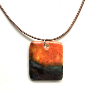 Puede incluir: Un collar de cordón de cuero marrón con un colgante rectangular de cerámica. El colgante es naranja, amarillo, verde y negro con una textura moteada.
