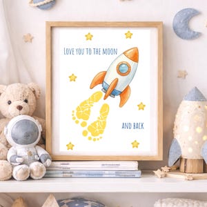 Könnte beinhalten: Ein gerahmter Druck mit einer Raketenillustration und dem Text "LOVE YOU TO THE MOON AND BACK". Die Rakete ist weiß und orange, mit gelben Fußabdrücken. Gelbe Sterne umgeben das Bild. Der Rahmen ist hellbraun.