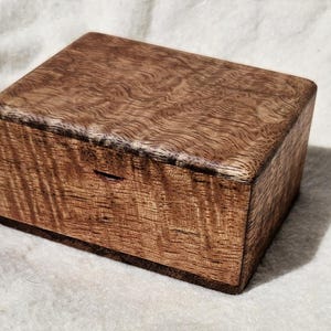 May include: A rectangular wooden box with a hinged lid, featuring a prominent wavy, rippled grain pattern in shades of medium to dark brown. The box has a smooth, polished finish and a small horizontal notch on the front for opening the lid.
