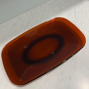 May include: An amber-coloured rectangular glass tray with a dark brown oval centre. The tray has a glossy finish and a slightly raised edge. The design is simple and modern, suitable for serving or display.