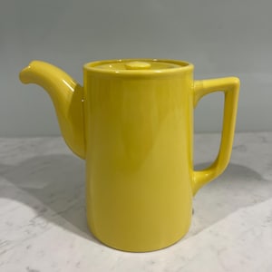 May include: A bright yellow ceramic teapot with a cylindrical body, a matching lid with a small central knob, a curved spout, and a rectangular handle. The teapot features a smooth, glossy finish and a solid, vibrant yellow colour throughout its exterior.