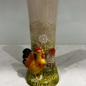 May include: A cylindrical ceramic vase featuring a raised, three-dimensional cockerel in shades of yellow, orange, and black. The background depicts a moulded farm scene with a white windmill and a small barn with a red roof against a beige sky.