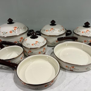 May include: A set of cream-coloured enamel cookware featuring a floral pattern of orange flowers and green leaves. The set includes various pots with matching lids and two skillets, all finished with dark brown handles and rim accents.