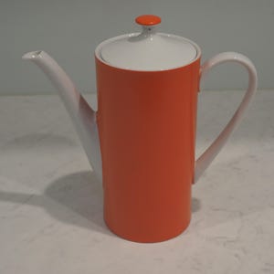 May include: A tall cylindrical teapot featuring a vibrant orange body with a contrasting white handle, spout, and lid. The lid is topped with a small, round orange knob. The design is sleek and vertical, showcasing a clean colour-blocked aesthetic.