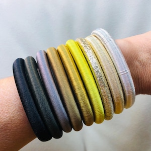 Può includere: Una collezione di braccialetti elastici colorati in varie tonalità, tra cui nero, grigio, oro e argento. I braccialetti sono impilati, mostrando il loro design strutturato e arrotolato. Un accessorio elegante.