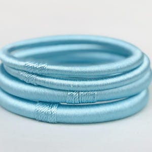 Könnte beinhalten: Ein Set aus vier hellblauen Stoffarmbändern. Die Armbänder sind aus einem weichen, gewebten Material gefertigt und übereinander gestapelt.