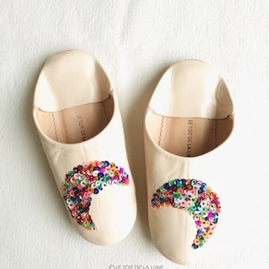 May include: A pair of cream-colored leather slippers with a crescent moon design made of colorful sequins. The slippers have the text "LE TOIT DE LA LUNE" on the inside.