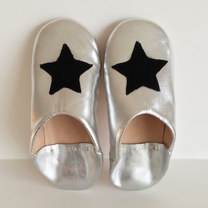 May include: A pair of silver leather slippers with black star appliques. The slippers have a pointed toe and a flat sole.
