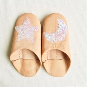 Puede incluir: Un par de zapatillas de cuero beige con un diseño de estrella y media luna hecho de lentejuelas iridiscentes. Las zapatillas son de dedos abiertos y tienen un diseño simple.