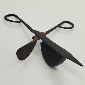 May include: An antique, rusty metal fire tool with a scissor-like design. The tool has two loop handles and a curved scoop on one side, with a flat, shovel-like shape on the other. The tool is likely used for handling hot coals.