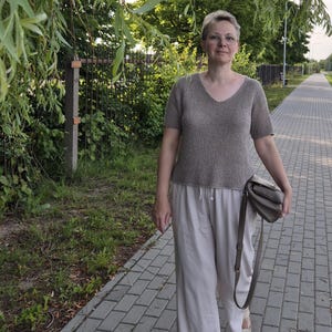 May include: A woman wearing a short-sleeved, V-neck brown top and light-colored pants walks on a brick path. She carries a gray purse with a shoulder strap and wears sandals. The background includes trees and a fence.