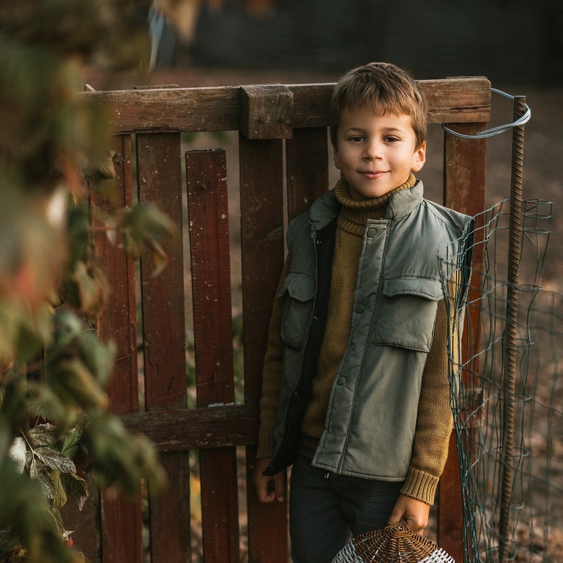 May include: A young boy wearing a green vest over a brown turtleneck sweater stands in front of a wooden fence. He is looking at the camera with a slight smile.