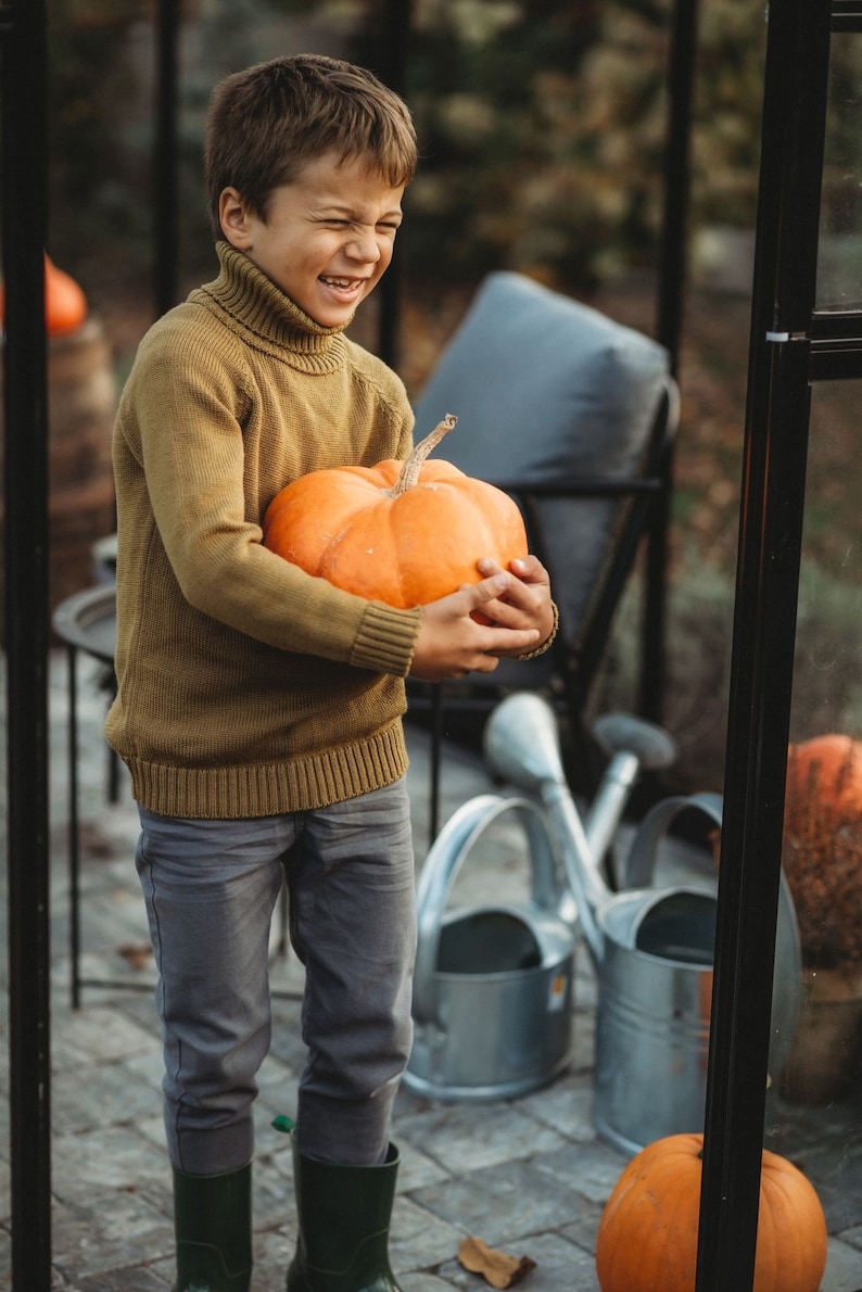 May include: A young boy wearing a brown sweater and grey pants holds a large orange pumpkin. He is smiling and looking at the camera.