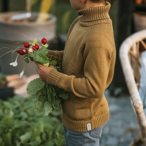 May include: A young person wearing a brown turtleneck sweater and blue jeans holds a bunch of fresh radishes with green leaves.