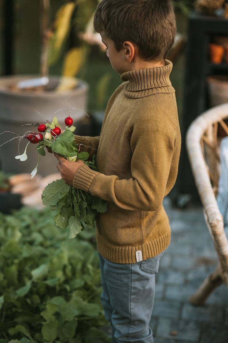 May include: A young person wearing a brown turtleneck sweater and blue jeans holds a bunch of fresh radishes with green leaves.