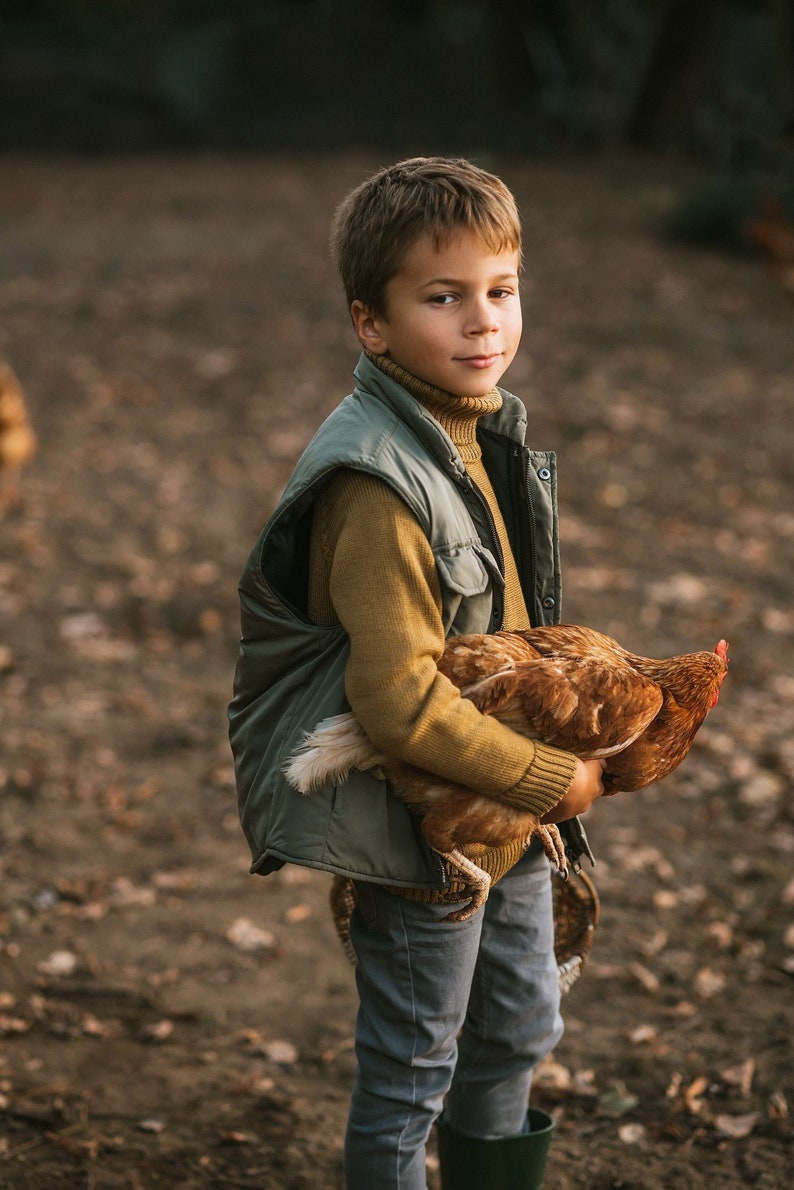 May include: A young person wearing a green vest and brown sweater holds a brown chicken in their arms. The person is standing in a dirt area with leaves and branches on the ground.