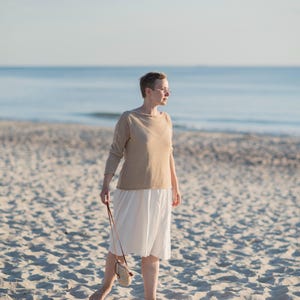 May include: A woman wearing a beige long-sleeve top and a white skirt stands on a sandy beach. She holds a small, round, beige purse with a brown strap. The background features the ocean and a clear sky.