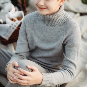 May include: A young person wearing a gray turtleneck sweater and gray pants sits on a white blanket. The person is looking off to the side and smiling.