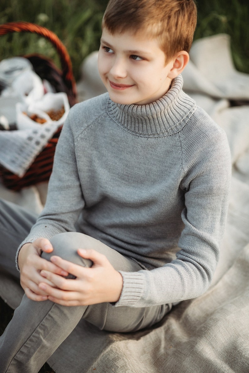 May include: A young person wearing a gray turtleneck sweater and gray pants sits on a white blanket. The person is looking off to the side and smiling.