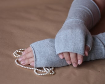 May include: A pair of gray fingerless gloves made of soft knit fabric. The gloves are folded and placed on a brown surface with a string of white pearls.