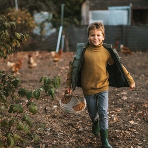 May include: A young boy in a brown turtleneck sweater and a green vest walks through a farm with a wicker basket in his hand. He is smiling and looking at the camera.