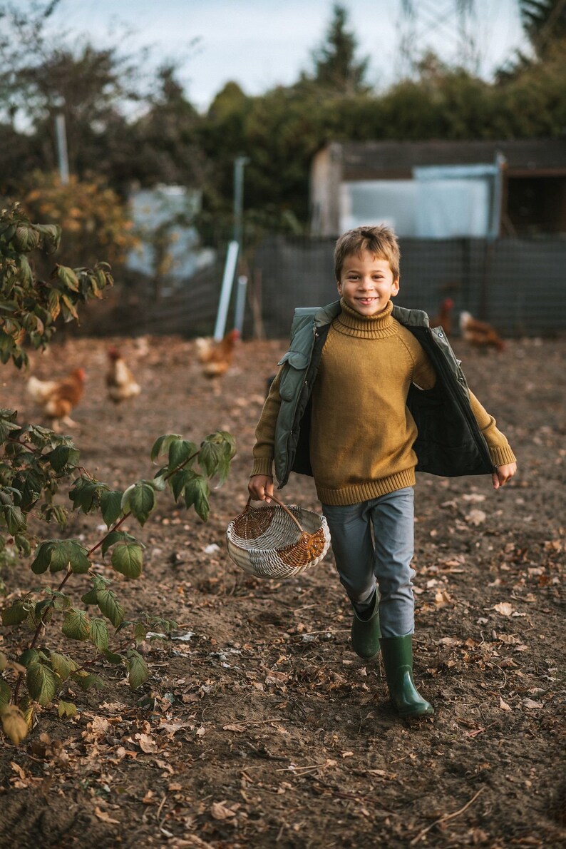 May include: A young boy in a brown turtleneck sweater and a green vest walks through a farm with a wicker basket in his hand. He is smiling and looking at the camera.
