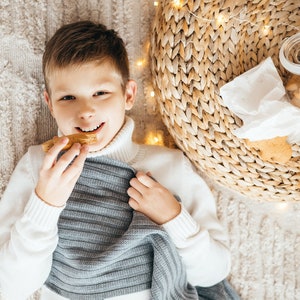 May include: A young boy wearing a white sweater and a gray knitted scarf is lying on a beige carpet. He is holding a piece of bread in his hand and smiling. There are string lights and a wicker basket in the background.