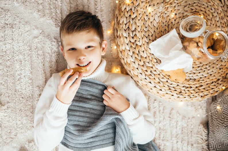 May include: A young boy wearing a white sweater and a gray knitted scarf is lying on a beige carpet. He is holding a piece of bread in his hand and smiling. There are string lights and a wicker basket in the background.
