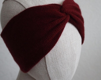 Puede incluir: Una diadema de punto en color burdeos intenso con un detalle de nudo retorcido en el centro. El accesorio está confeccionado con un hilo suave y texturizado, y se muestra sobre una cabeza de maniquí de color neutro.