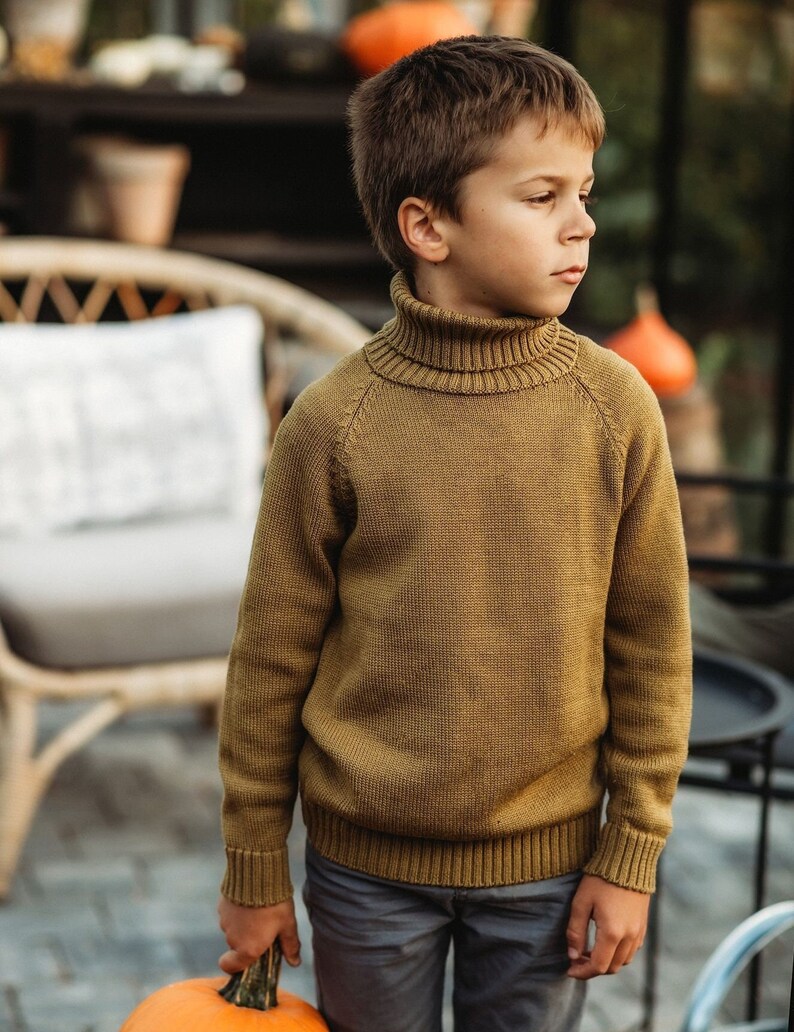 May include: A young boy wearing a brown turtleneck sweater and gray pants holds a large orange pumpkin.