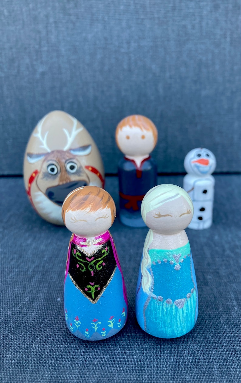May include: A set of five wooden peg dolls, including a reindeer, a boy with brown hair, a girl with blonde hair, a girl with brown hair, and a snowman. The dolls are painted in various colours, including blue, green, pink, and white.