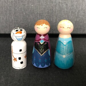 May include: A set of three hand-painted wooden peg dolls featuring characters from the film Frozen. The dolls are Olaf the snowman, Elsa, and Anna. Olaf is white with a carrot nose and black eyes. Elsa is wearing a blue dress with a sparkly bodice. Anna is wearing a purple dress with a black and gold bodice.
