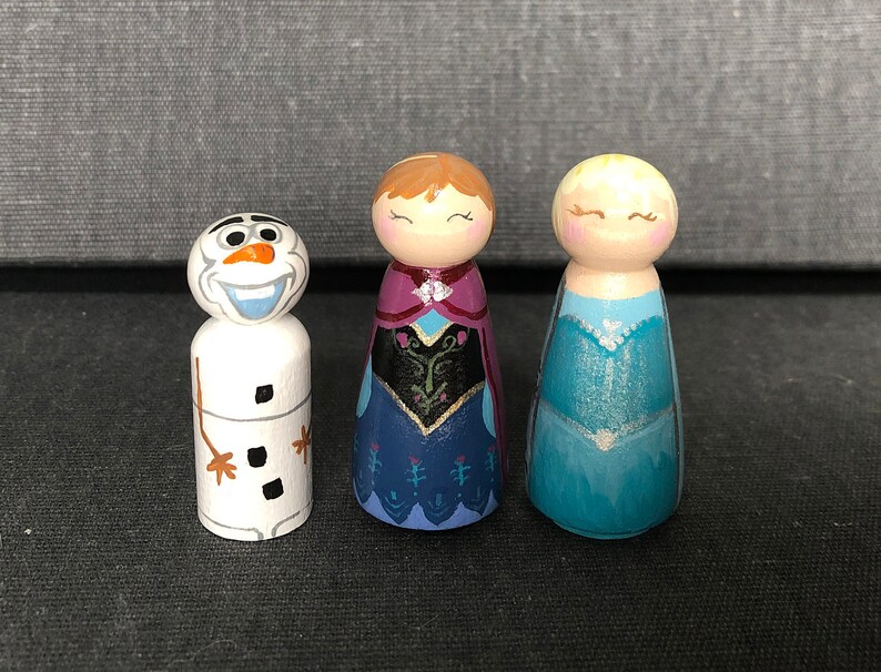 May include: A set of three hand-painted wooden peg dolls featuring characters from the film Frozen. The dolls are Olaf the snowman, Elsa, and Anna. Olaf is white with a carrot nose and black eyes. Elsa is wearing a blue dress with a sparkly bodice. Anna is wearing a purple dress with a black and gold bodice.