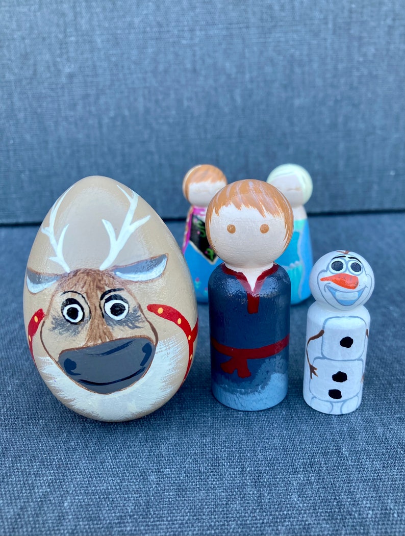 May include: A set of four hand-painted wooden figurines featuring characters from the film Frozen. The set includes a reindeer, a boy with brown hair, a girl with blonde hair, and Olaf the snowman. The figurines are painted in bright colours and have a whimsical style.