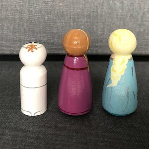 May include: Three wooden peg dolls, one with a white face and brown hair, one with a purple dress and brown hair, and one with a blue dress and blonde hair.