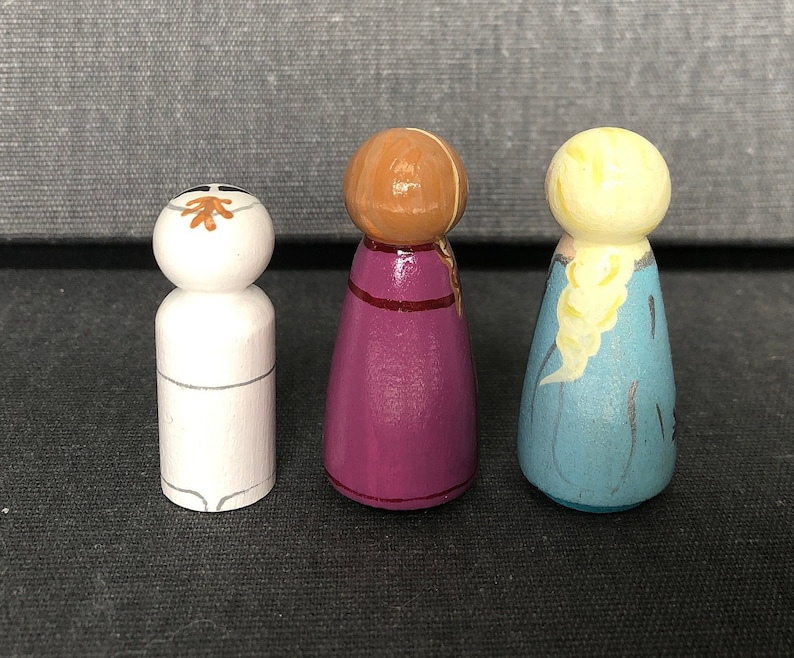 May include: Three wooden peg dolls, one with a white face and brown hair, one with a purple dress and brown hair, and one with a blue dress and blonde hair.