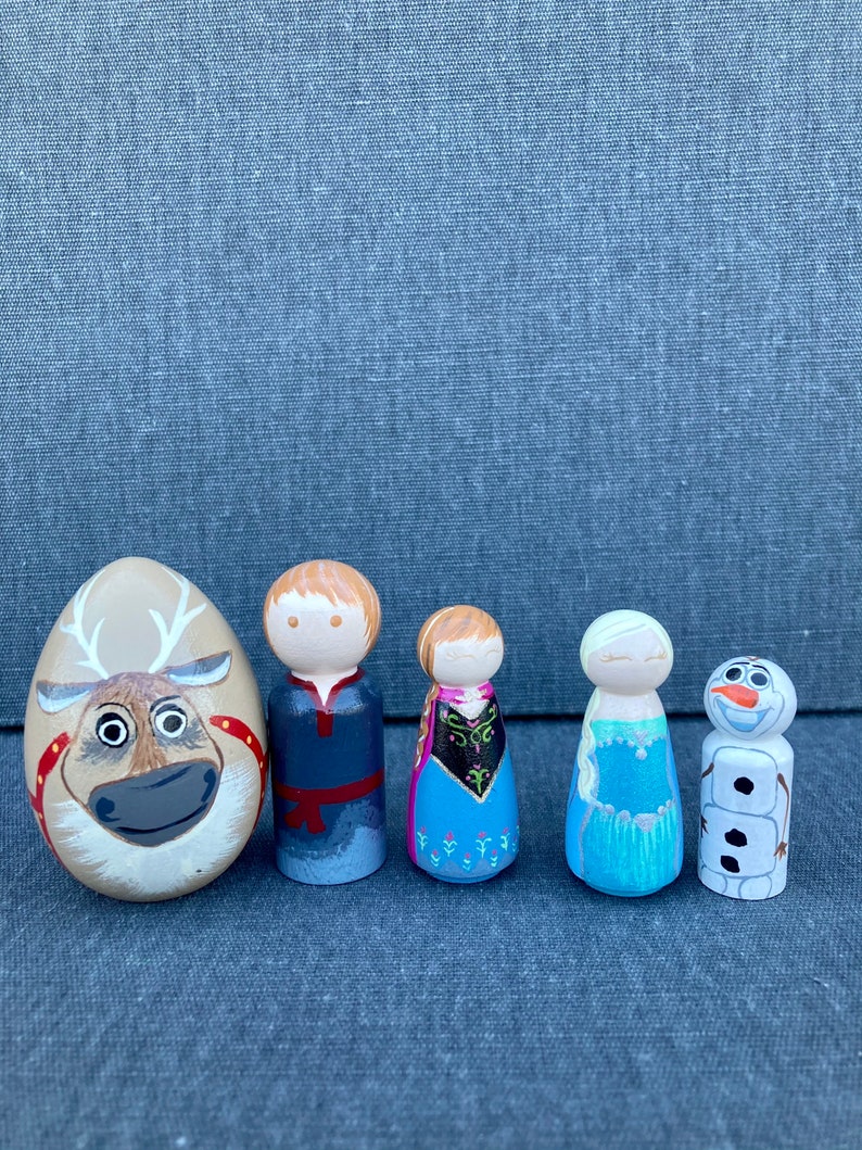 May include: A set of five wooden peg dolls featuring characters from the animated film Frozen. The dolls are painted in the likeness of Elsa, Anna, Kristoff, Olaf, and Sven the reindeer.