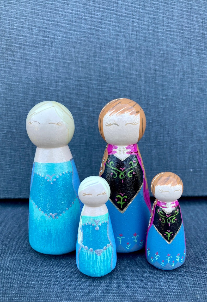 May include: Set of four wooden peg dolls dressed as characters from the film Frozen. The dolls are painted in blue, pink, and black with glitter accents. The dolls have brown and blonde hair and closed eyes.