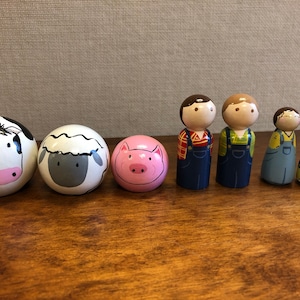 May include: A set of wooden peg dolls depicting a farm scene. The set includes a cow, sheep, pig, two farmers, and a girl. The dolls are painted in bright colors and have simple facial features.