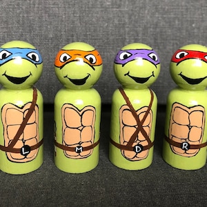 May include: Four wooden peg dolls painted to look like the Teenage Mutant Ninja Turtles. Each doll has a different colored mask and a unique letter on its belt. The dolls are green with orange, purple, blue, and red masks.