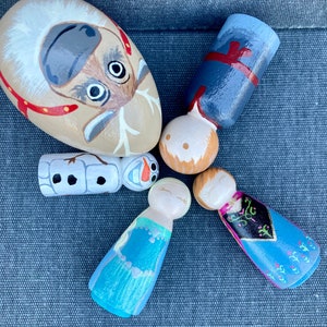 May include: A set of five hand-painted wooden figurines depicting characters from the film Frozen. The figurines are painted in various shades of blue, purple, and white. The set also includes a painted rock depicting Sven the reindeer.