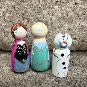 May include: Three hand-painted wooden peg dolls featuring characters with distinct outfits. One doll has brown hair and a black and pink dress, another has blonde hair and a light blue dress, and the third is a white snowman with a carrot nose.