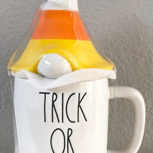 May include: White ceramic mug with the words "TRICK OR TREAT" in black lettering. The mug is topped with a candy corn-shaped lid in orange, yellow, and white. A handle is attached to the side of the mug.