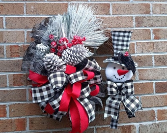 May include: A black and white buffalo plaid Christmas wreath with a red bow and a snowman ornament. The wreath is decorated with pine cones, berries, and faux snow.